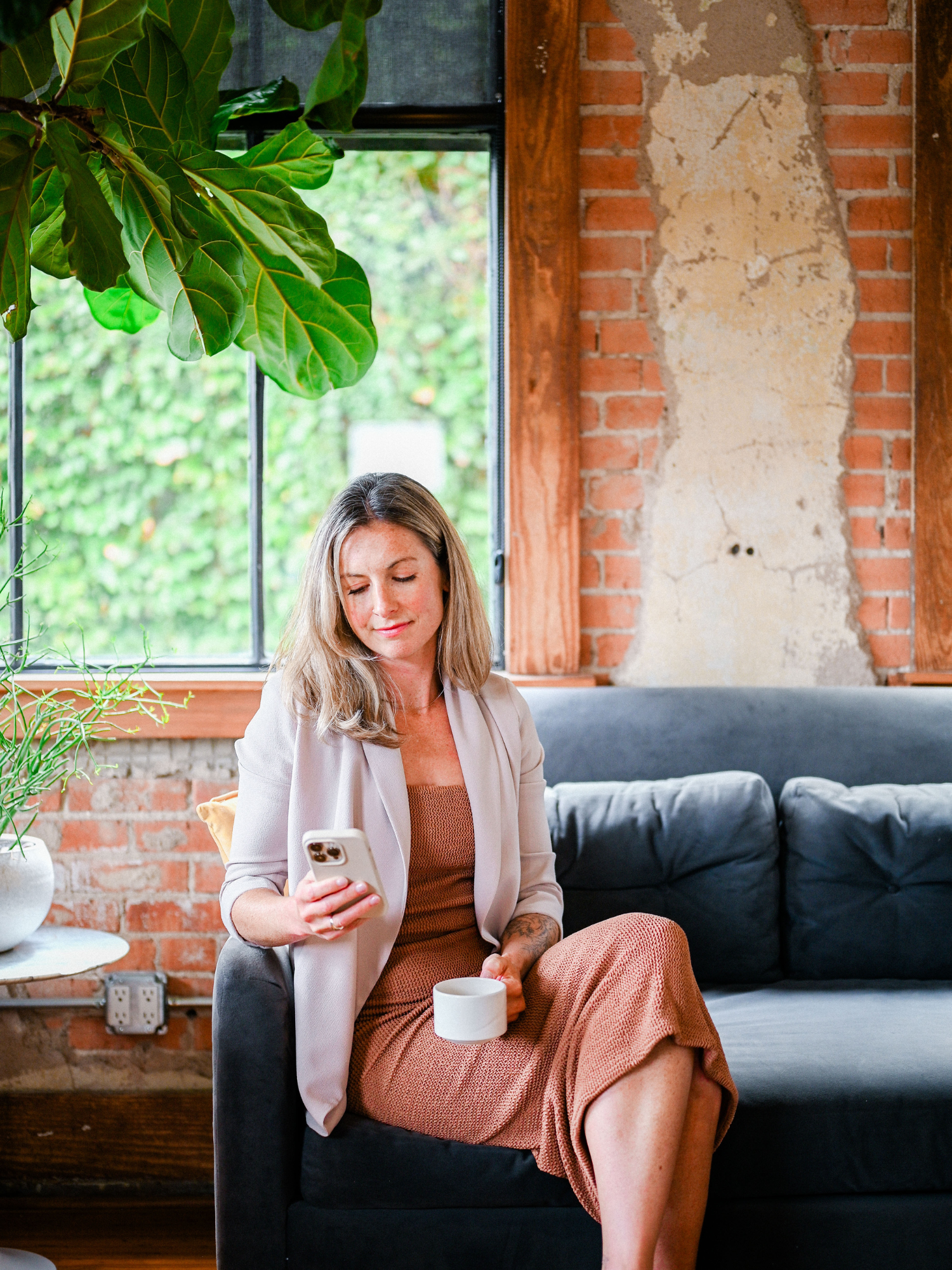 Jaime Bell, Lawyer sitting on a coach reading her phone. Wearing a brown dress and beige jacket, tree in background with brick wall